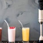Three smoothies in glasses with straws—chocolate, pink with raspberries, and yellow—are lined up next to an immersion blender against a gray background.