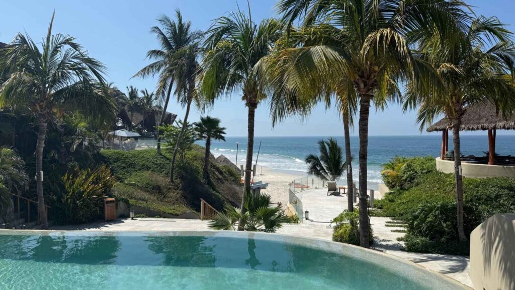 A clear swimming pool at one of the finest family wellness resorts overlooks palm trees, a sandy beach, and the ocean with gentle waves under a sunny blue sky.