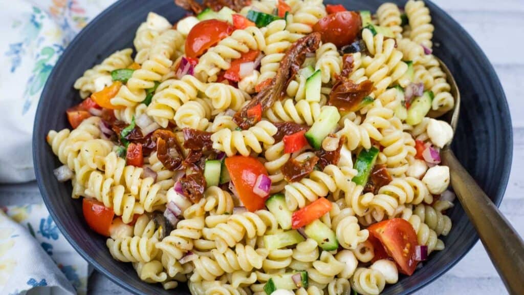 A bowl of Italian pasta salad with cherry tomatoes, cucumber, red onion, mozzarella balls, and sun-dried tomatoes, served with a fork.