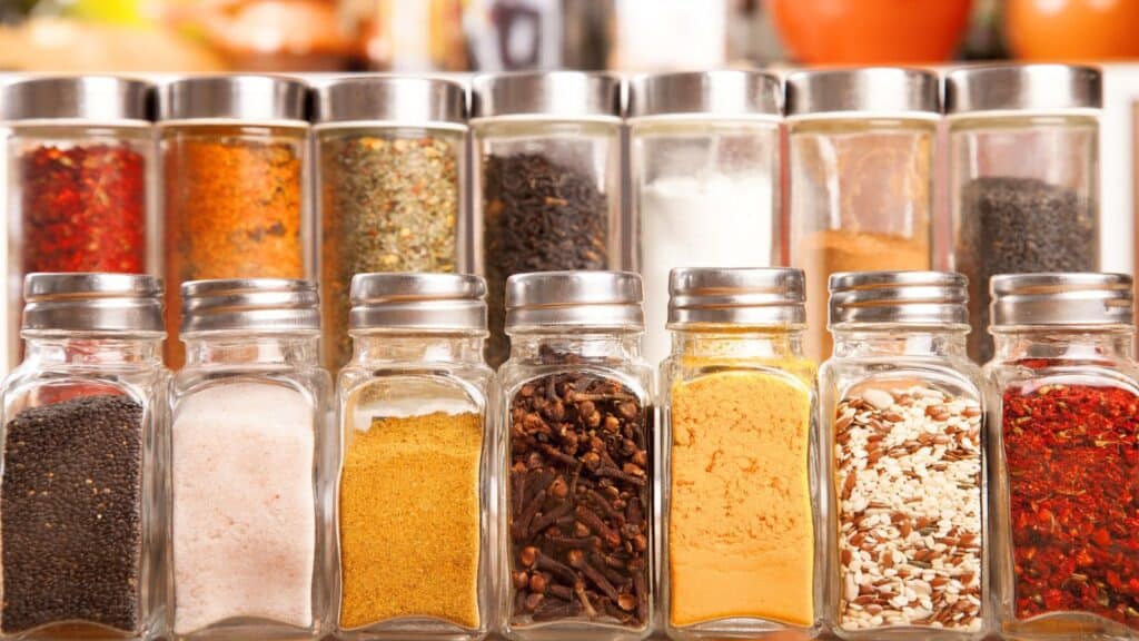 A row of glass spice jars with metal lids, each containing different colorful spices and seasonings, arranged on a kitchen counter.