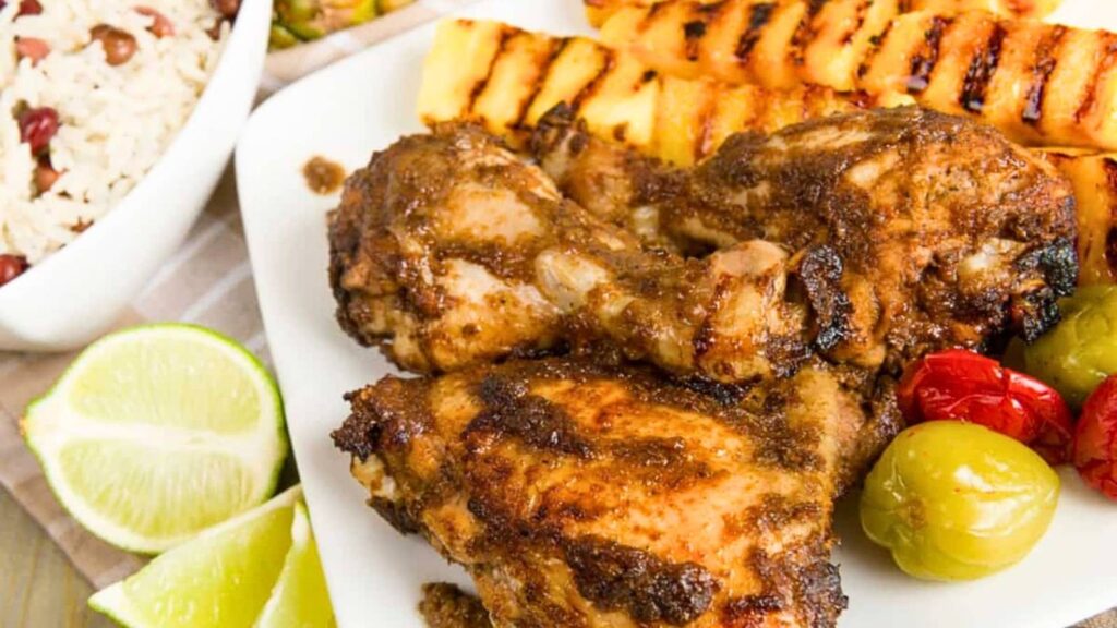 Grilled jerk chicken drumsticks on a plate with grilled pineapple, roasted peppers, and lime wedges, served alongside a bowl of rice and peas.