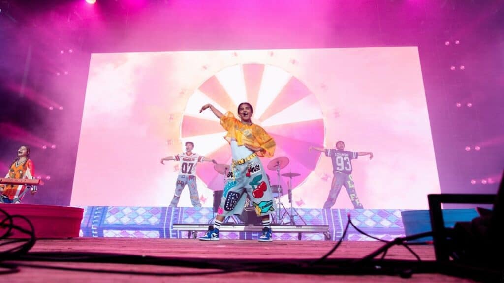 A group of performers in colorful outfits dance and play instruments on a stage with a bright, graphic backdrop and pink lighting.