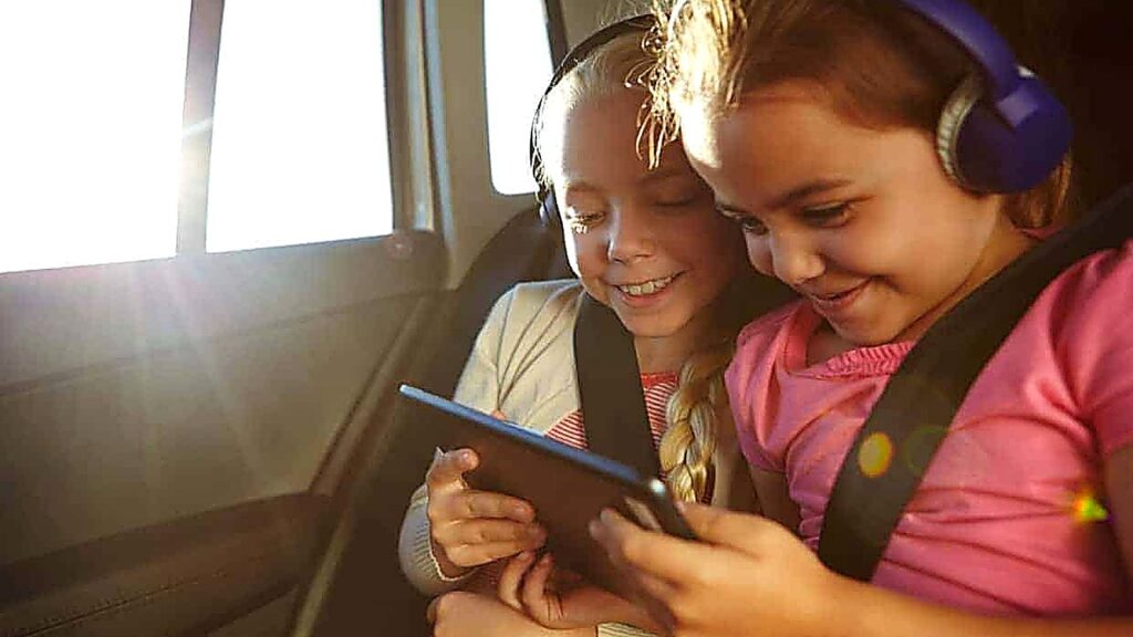 Two young children in car seats wearing headphones and watching a tablet together in the backseat of a car. Sunlight comes through the window.