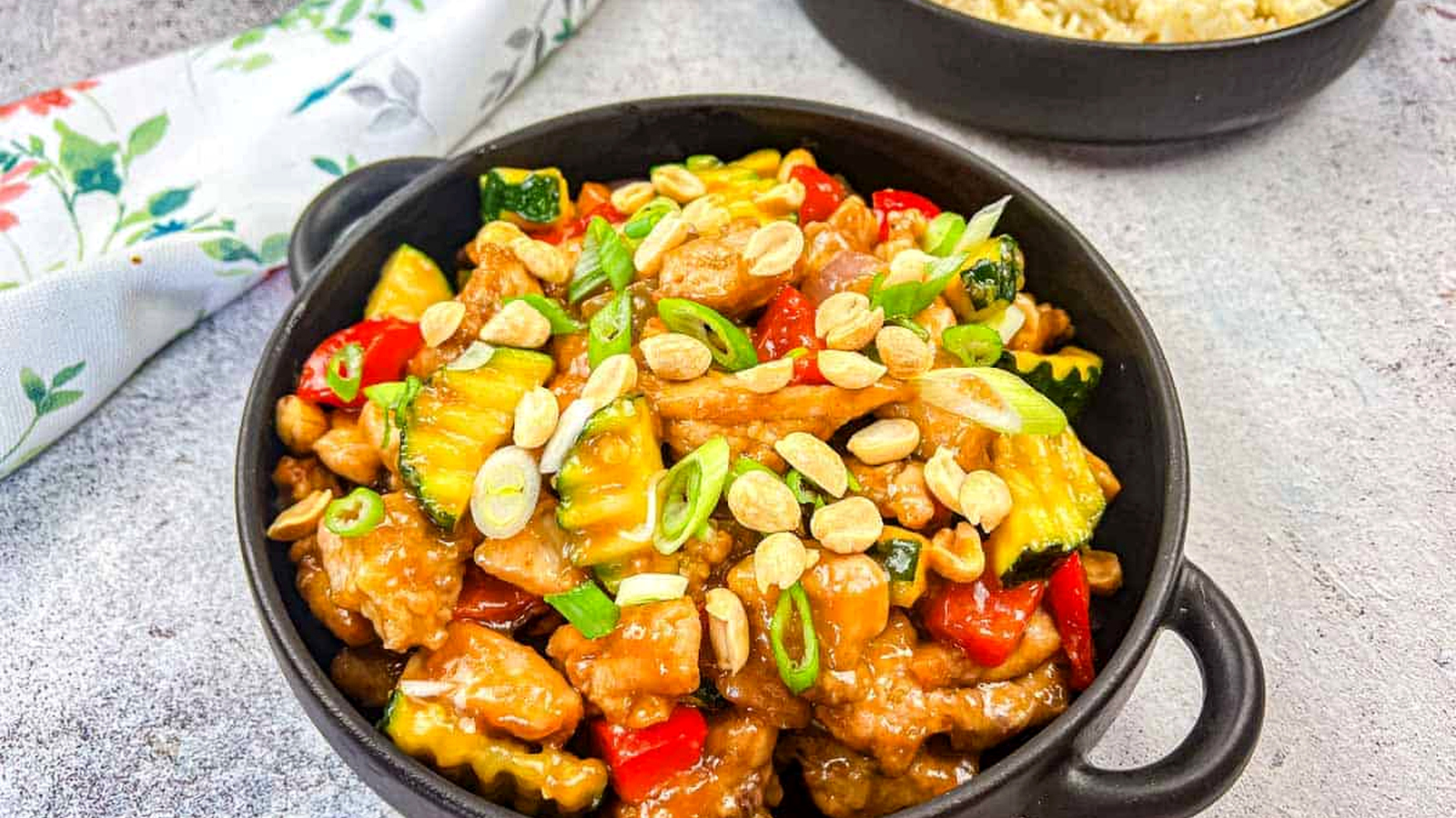 Kung Pao Chicken in a bowl with a bowl of rice in the background.