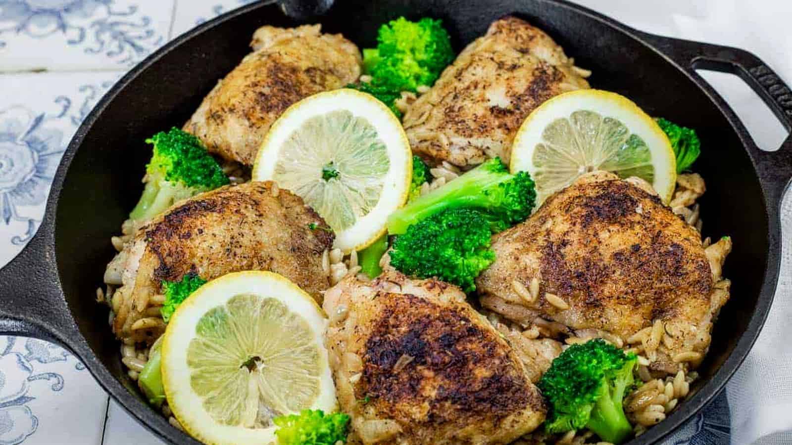 Golden-browned lemon chicken thighs served over orzo pasta with tender broccoli florets, garnished with fresh lemon slices in a cast-iron skillet.