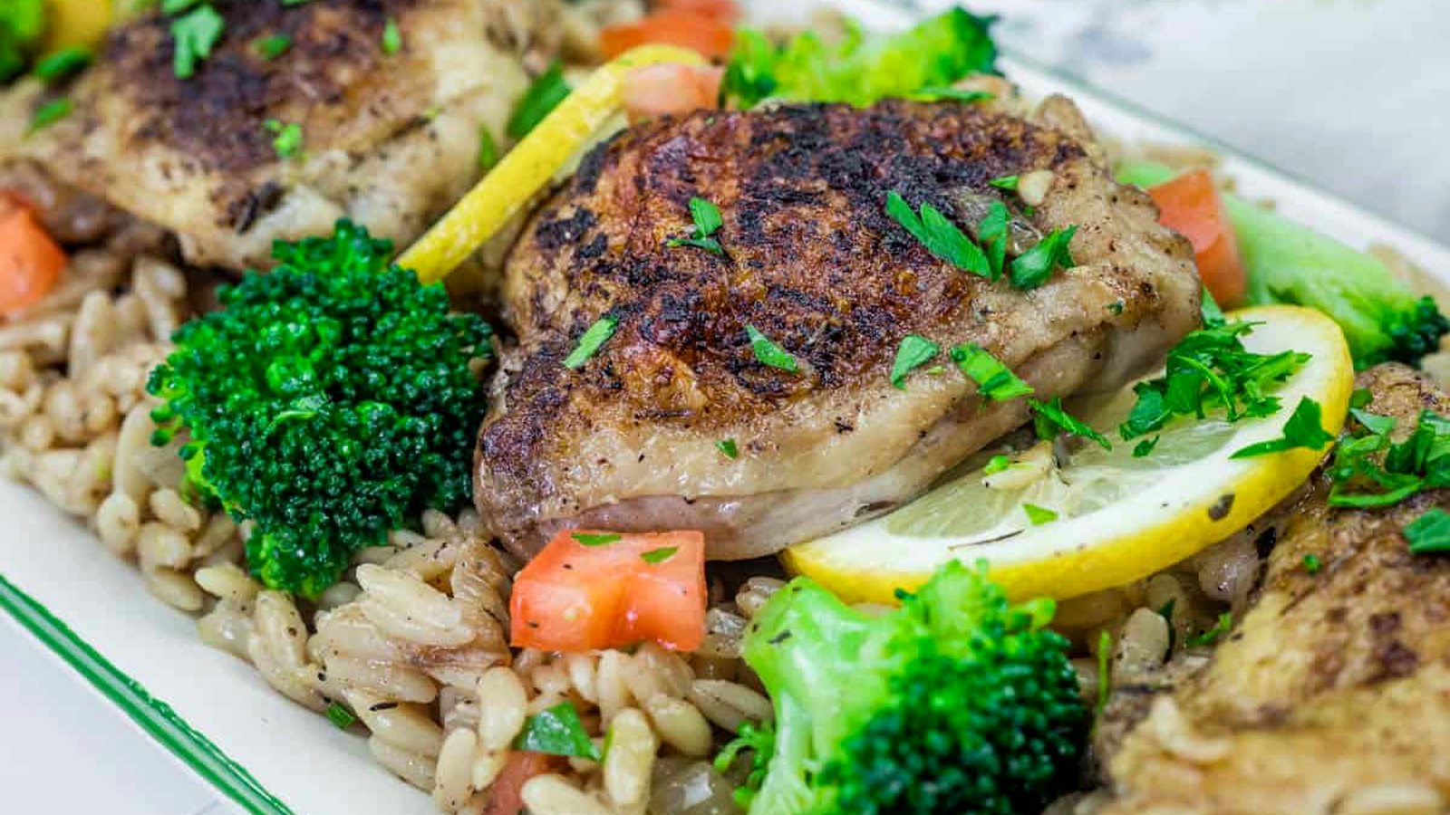 Golden-browned lemon chicken thighs served over orzo pasta with tender broccoli florets, garnished with fresh lemon slices in a cast-iron skillet.