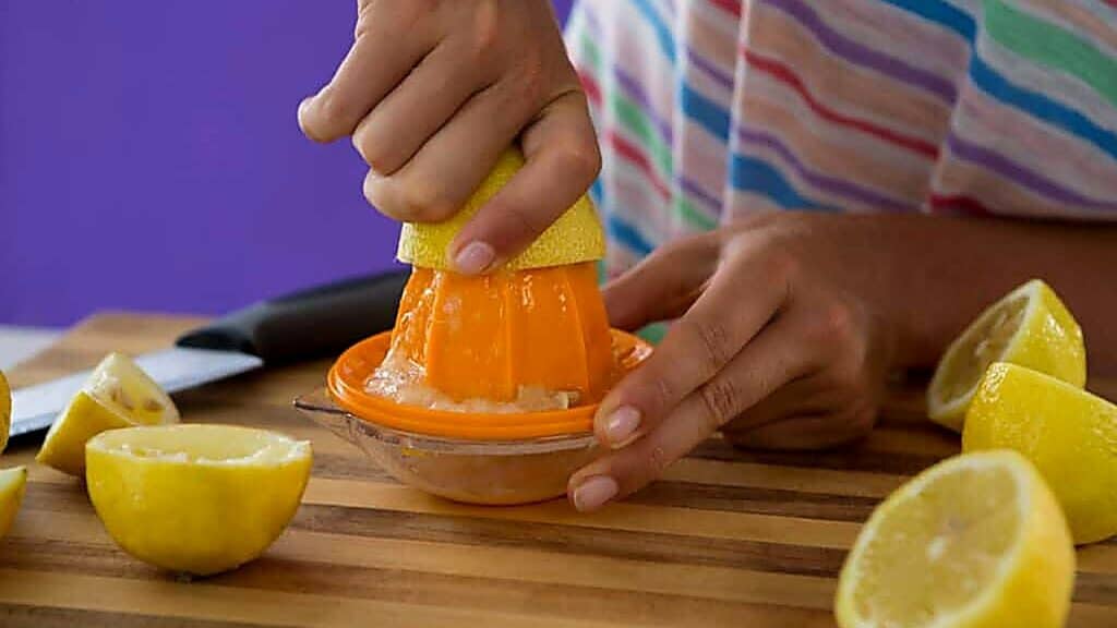 Person juicing a lemon with a hand citrus juicer on a wooden cutting board, surrounded by cut lemon halves.
