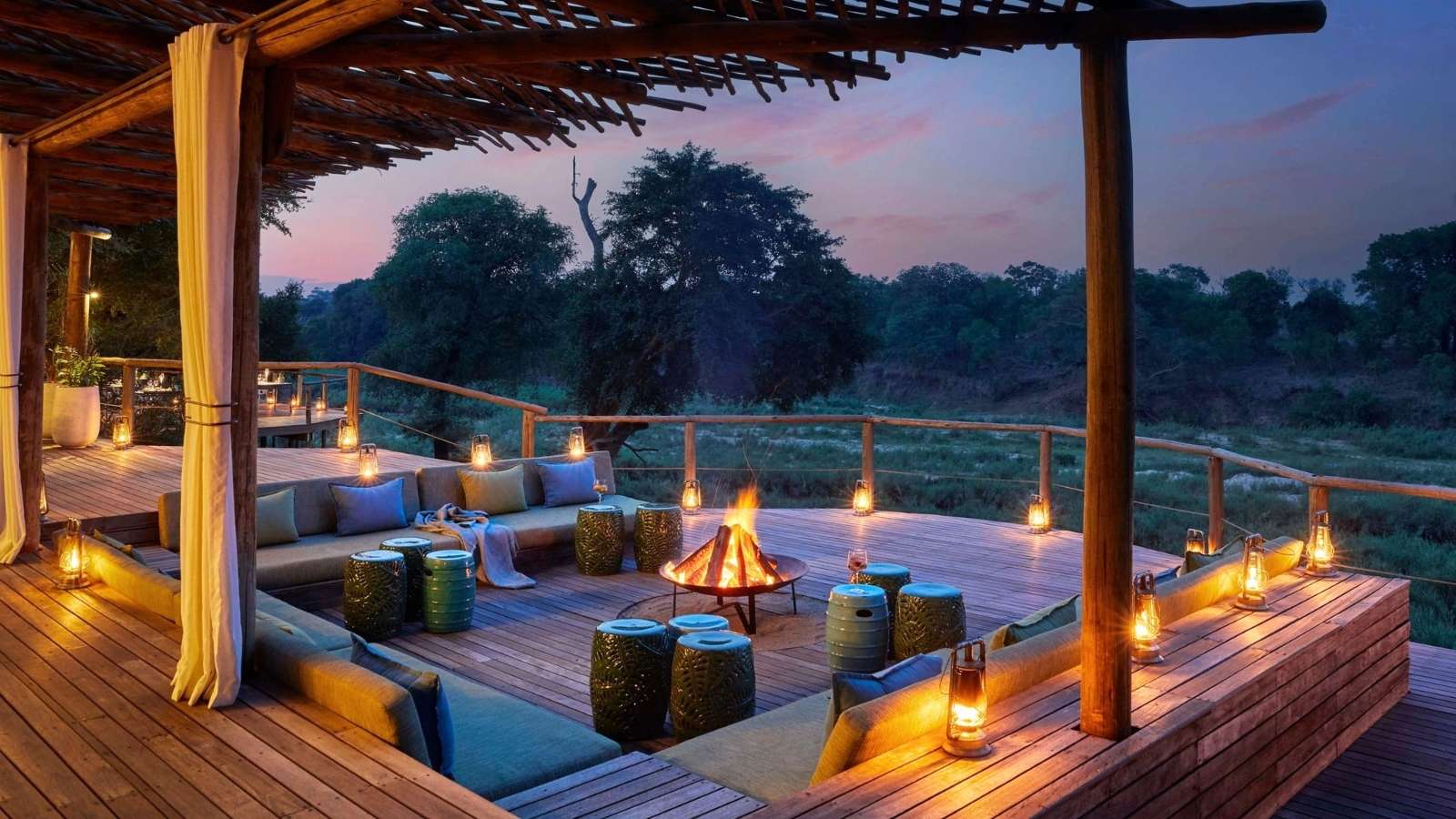 Outdoor wooden deck with cushioned seating and small tables around a fire pit, surrounded by lanterns, overlooking a scenic natural landscape at sunset.
