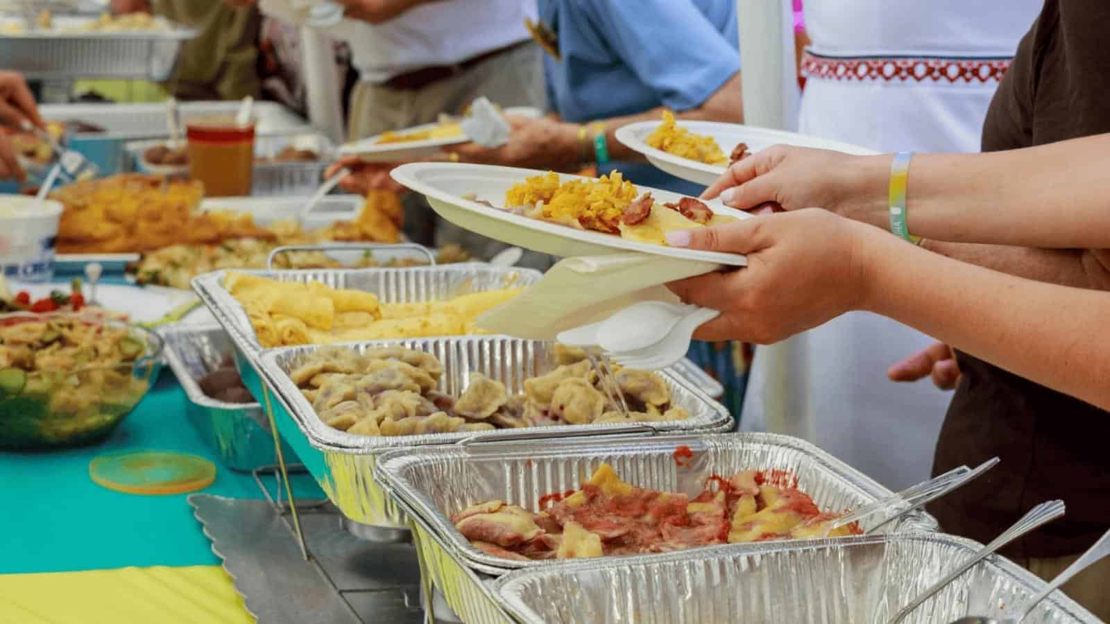 People serve themselves food from trays at a buffet table. Plates are filled with various dishes, and hands are visible holding plates and serving utensils.