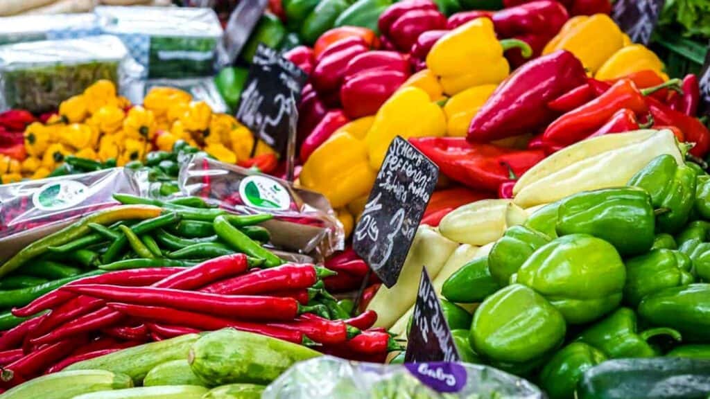 A colorful assortment of bell peppers and chili peppers displayed at a market stall, with handwritten price signs and packaged vegetables nearby, invites shoppers to gather fresh ingredients perfect for vegetable preparation.