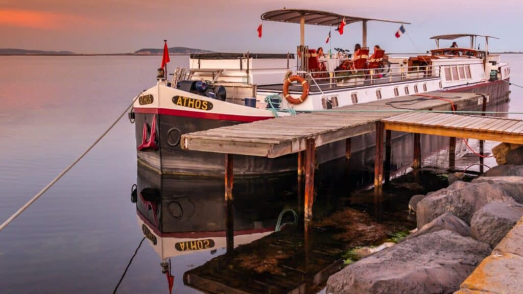 A white canal barge named "Athos" is moored beside a wooden dock on calm water at sunset, with reflections visible in the water.