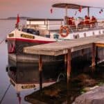 A white canal barge named "Athos" is moored beside a wooden dock on calm water at sunset, with reflections visible in the water.