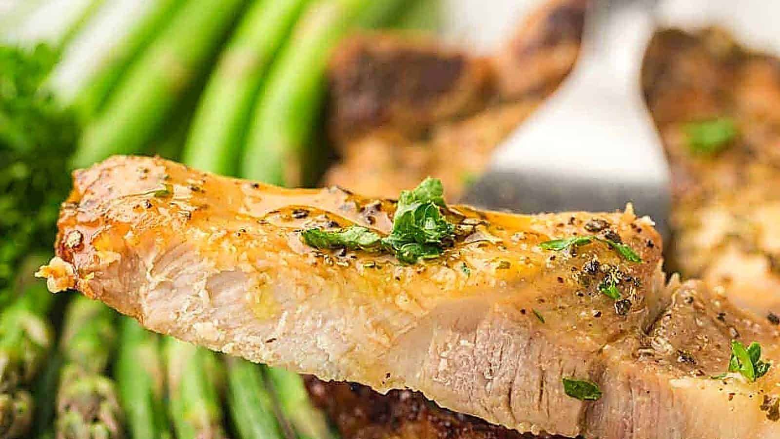 A close-up of a seasoned pork chop slice garnished with herbs, held by a fork, with asparagus spears in the background.