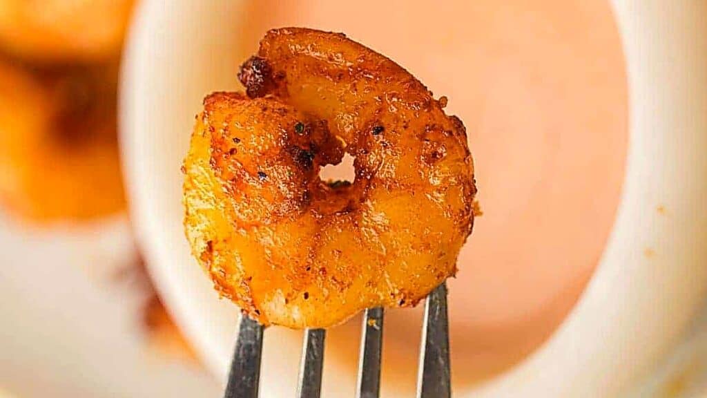 A cooked, seasoned shrimp held on a fork in front of a bowl of pink dipping sauce.