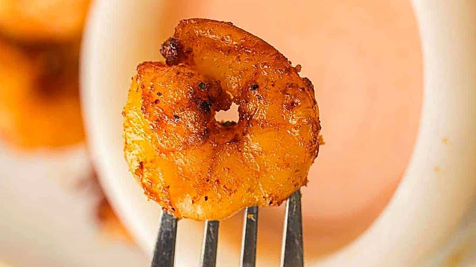 A close-up of a seasoned, cooked shrimp on a fork, held in front of a bowl of dipping sauce.