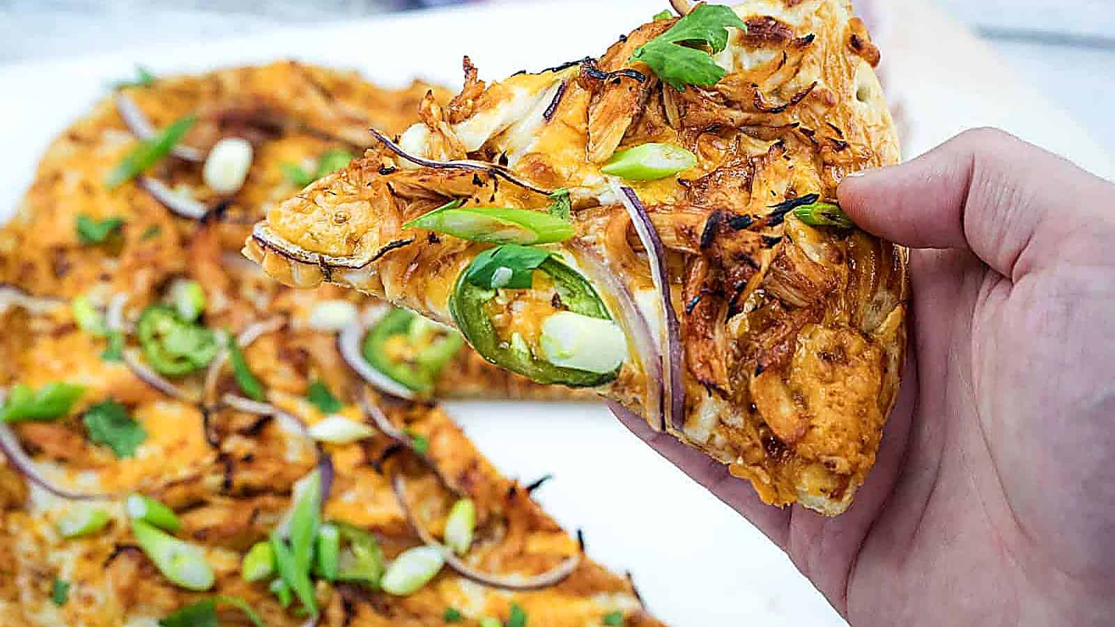 A hand holds a slice of pizza topped with shredded chicken, jalapeño slices, red onions, and green onions; the rest of the pizza is visible on a white surface.