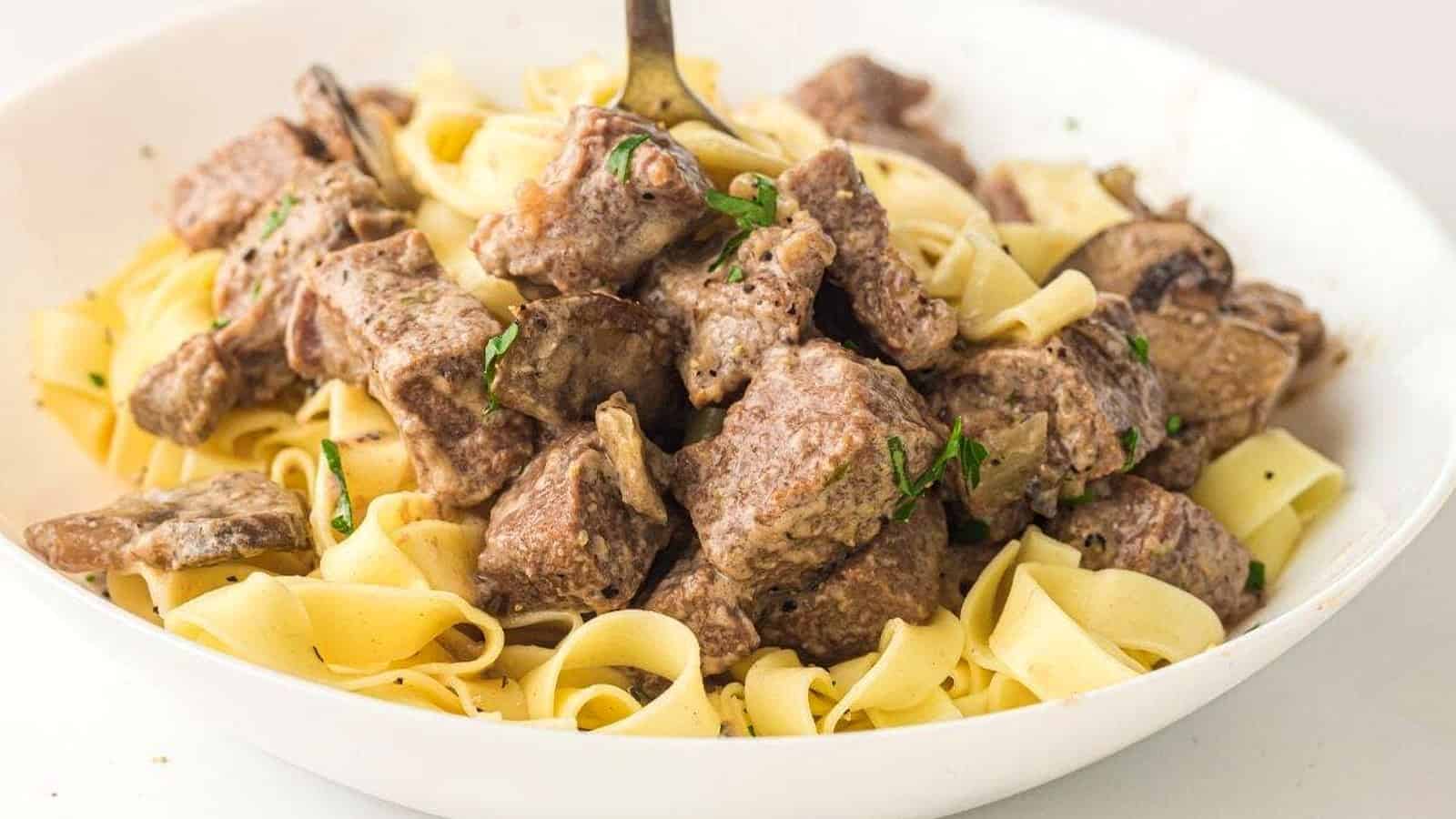 A white bowl filled with cooked egg noodles and chunks of beef, garnished with herbs, with a fork standing upright in the center.