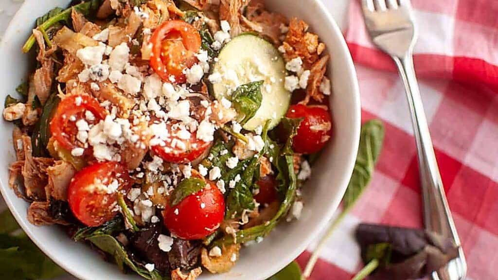 A bowl of salad with cherry tomatoes, cucumbers, greens, shredded meat, and crumbled cheese sits on a red-and-white checkered tablecloth beside a fork.