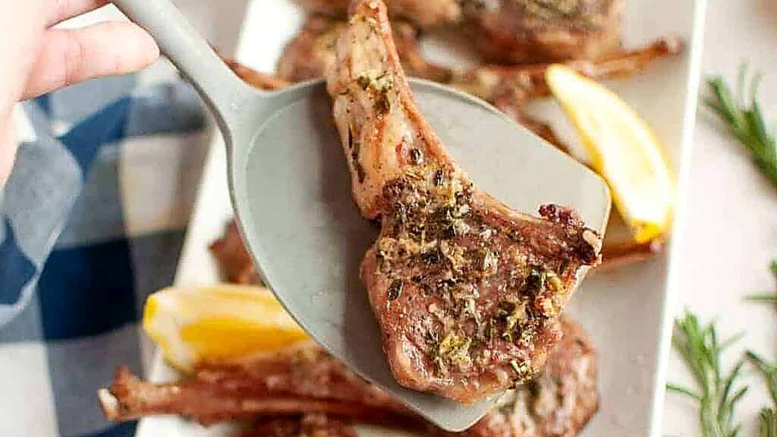 A cooked lamb chop with herbs is lifted by a spatula above a plate with more lamb chops and lemon wedges.