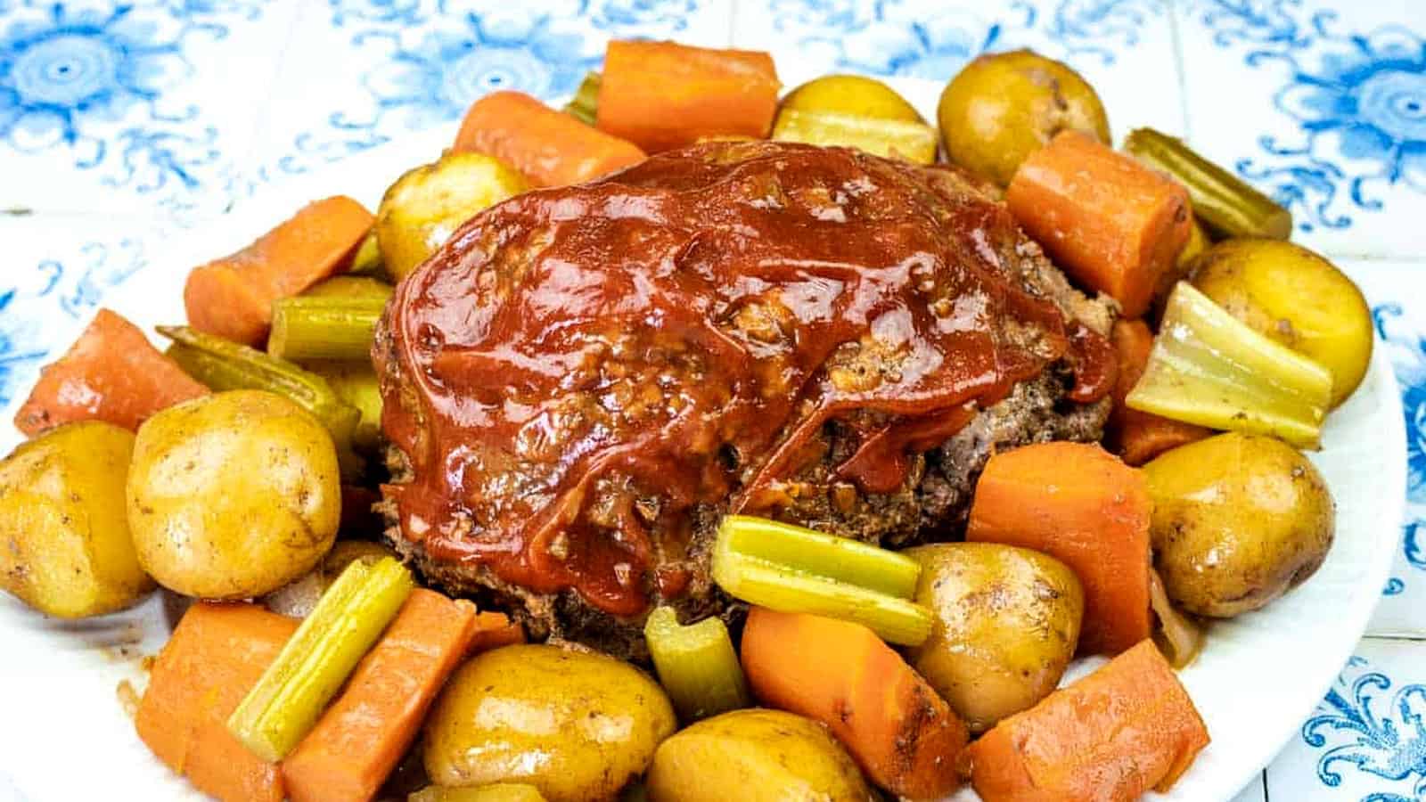 A plate of meatloaf topped with tomato sauce, surrounded by cooked baby potatoes, carrots, and celery on a patterned tile background.