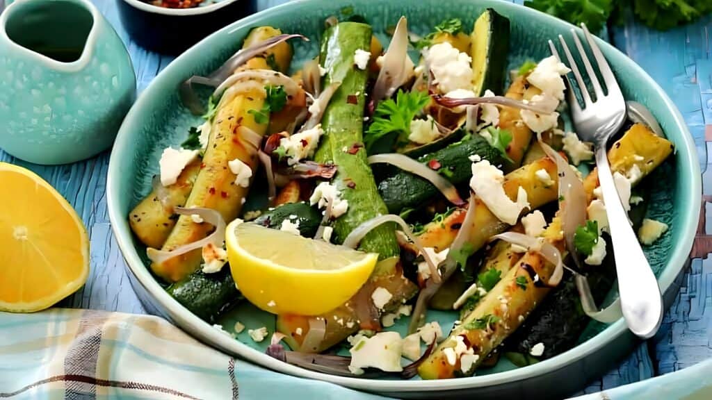 A plate of roasted zucchini with crumbled feta, sliced onions, parsley, and lemon wedges, served with a fork on a blue surface.