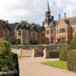 Large historic manor with red brick and timber detailing, tall chimneys, and a central clock tower, surrounded by manicured hedges and gardens under a partly cloudy sky.