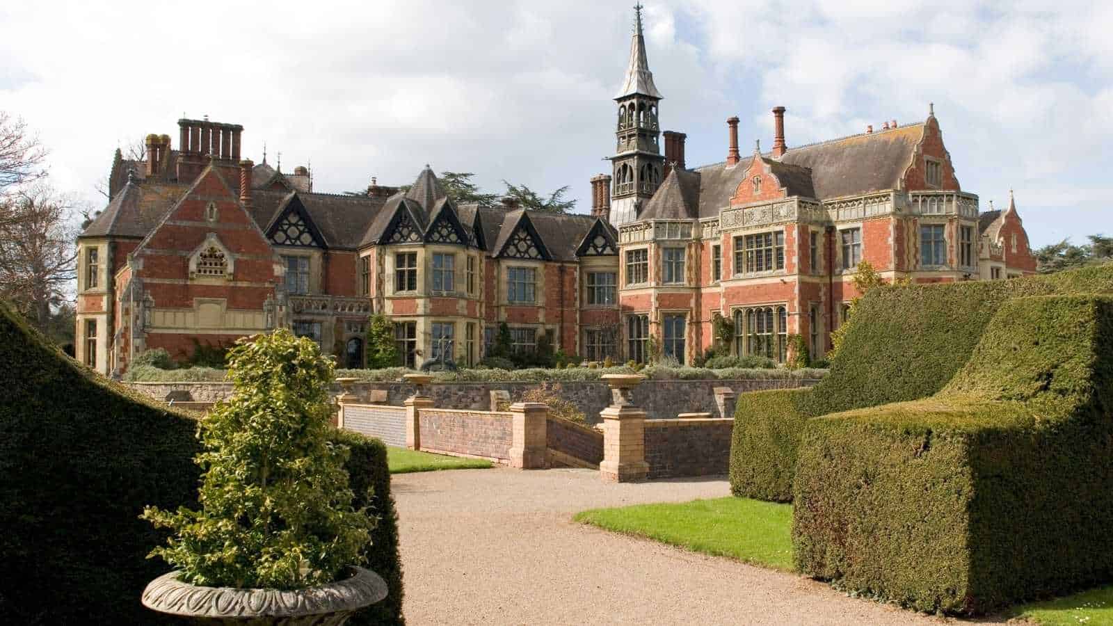 Large historic manor with red brick and timber detailing, tall chimneys, and a central clock tower, surrounded by manicured hedges and gardens under a partly cloudy sky.