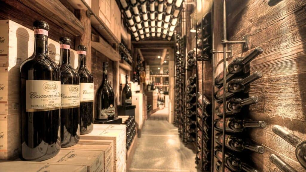 A wine cellar with shelves and racks filled with wine bottles in a dimly lit, rustic setting; bottles are stacked horizontally and vertically along the walls.