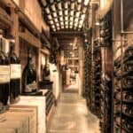 A wine cellar with shelves and racks filled with wine bottles in a dimly lit, rustic setting; bottles are stacked horizontally and vertically along the walls.