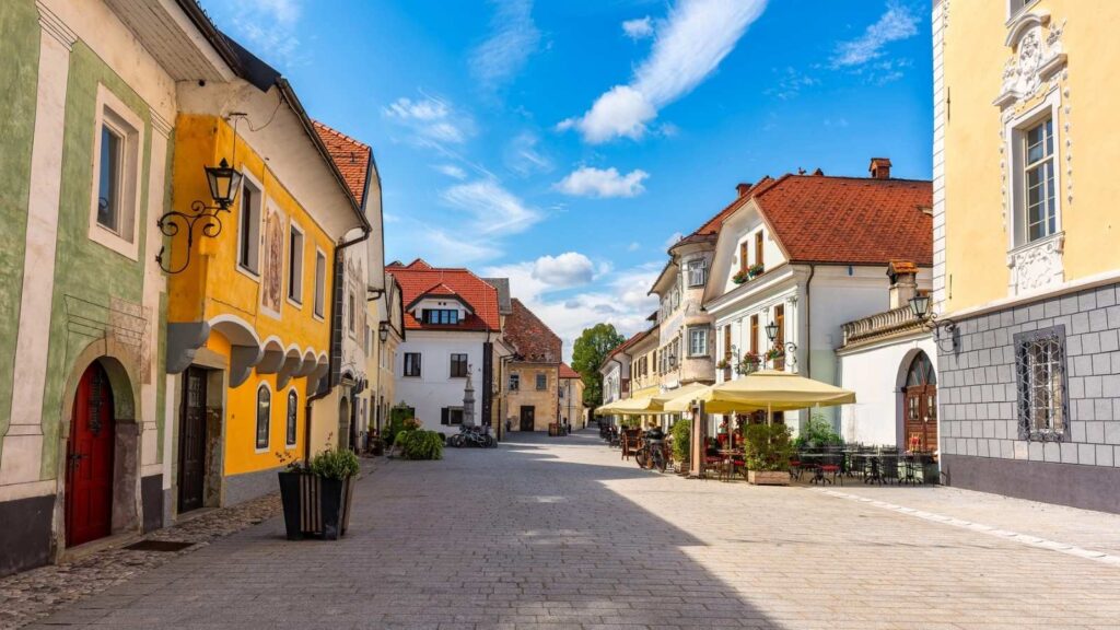 A cobblestone street lined with colorful historic buildings, outdoor cafes with yellow umbrellas, and a bright blue sky with scattered clouds set the scene as more Americans discover Slovenia&rsquo;s charming hidden gems.