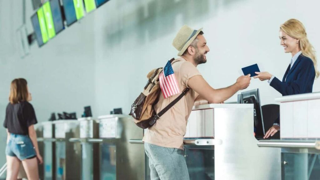 A man with a backpack hands his passport to an airport check-in agent at a counter, preparing for Fourth of July travel, while another woman walks by in the background.