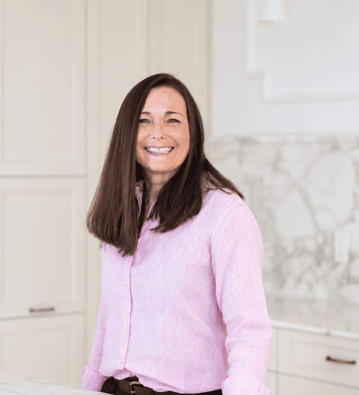 Woman with long brown hair, wearing a light pink button-up shirt, stands and smiles in a modern kitchen with light cabinets and a marble backsplash.