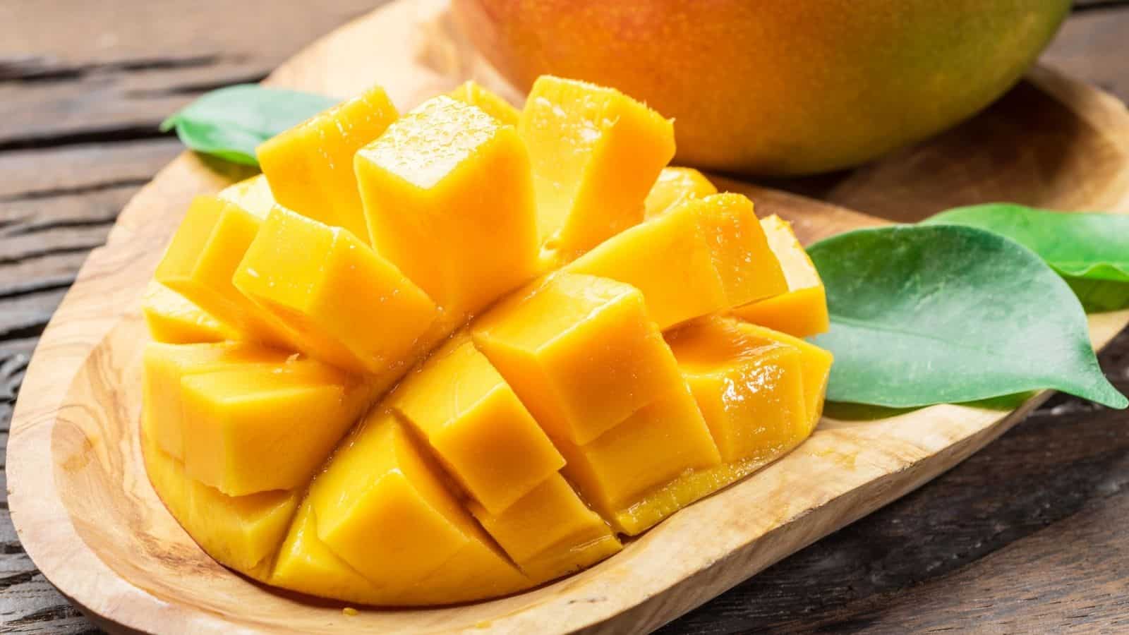 Cubed mango half displayed on a wooden plate with green leaves, with a whole mango in the background on a rustic wooden surface.