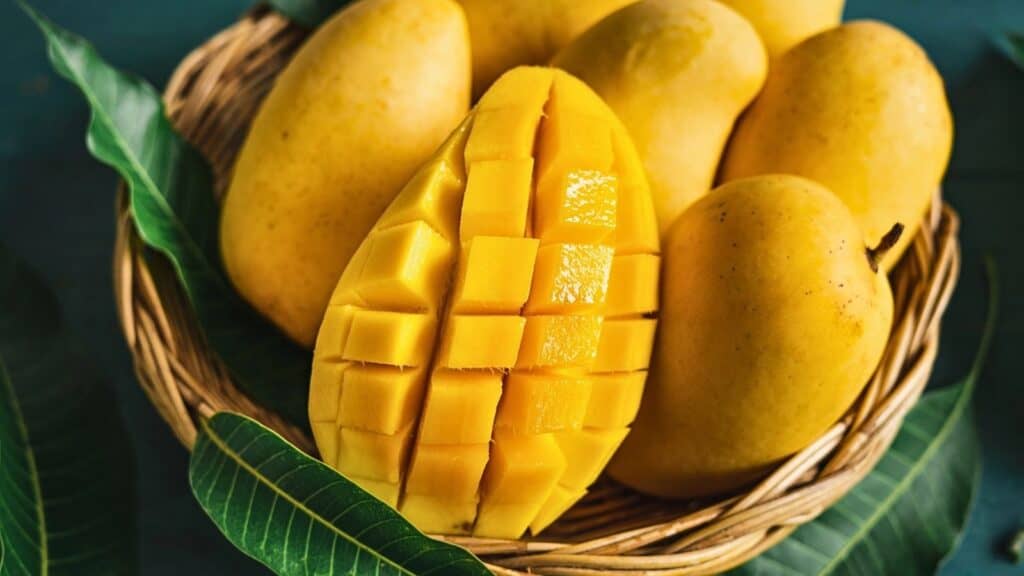 A wicker basket holds several whole yellow mangoes and one mango that is cut into cubes, with green mango leaves placed around the basket.