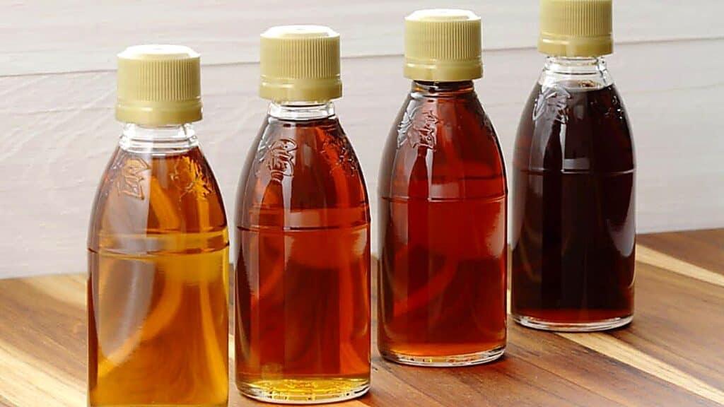 Four plastic bottles with beige caps filled with different shades of liquid, ranging from light amber to dark brown, are arranged in a row on a wooden surface.