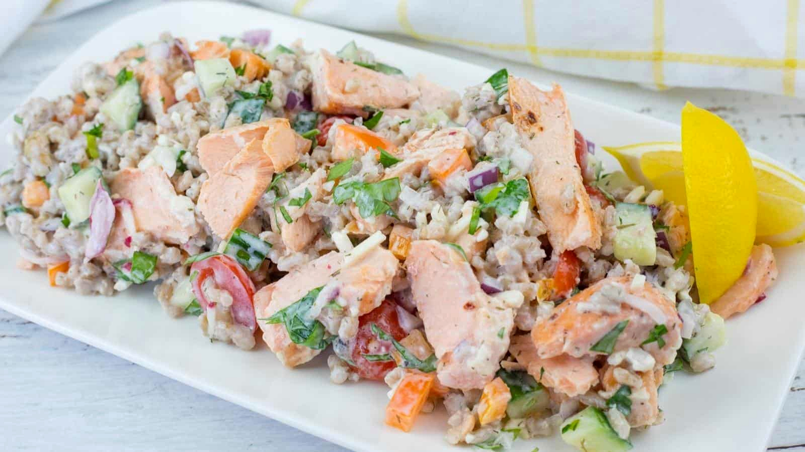 A rectangular white plate of Mediterranean Salmon Salad with Barley, topped with fresh herbs, and a lemon wedge on the side.