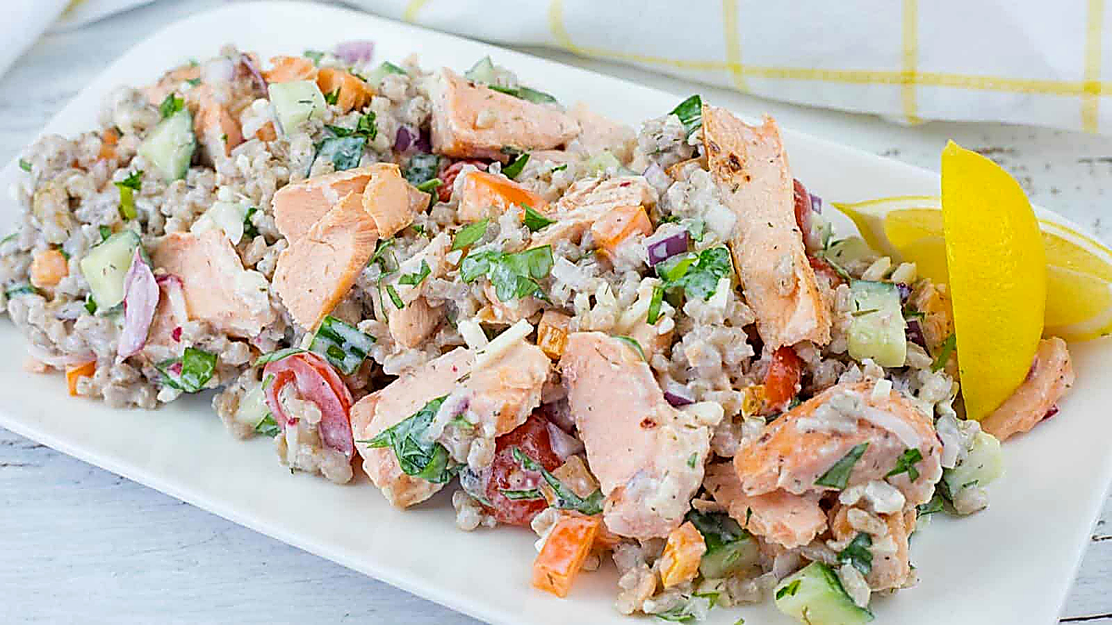 A rectangular white plate of Mediterranean Salmon Salad with Barley, topped with fresh herbs, and a lemon wedge on the side.