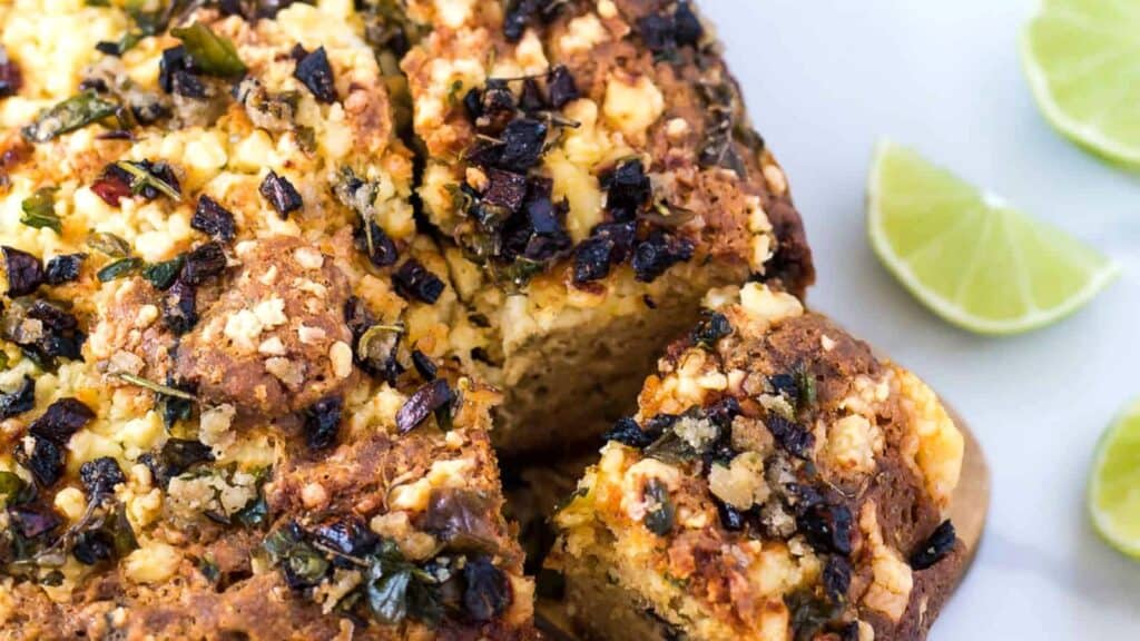 Close-up of a savory baked bread topped with crumbled cheese, herbs, and chopped vegetables, with lime wedges on the side.
