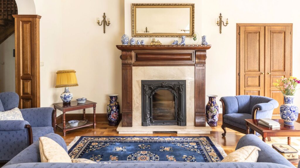A traditional living room with a fireplace, blue sofas, a blue rug, wooden furniture, vases, and a large mirror above the mantel.