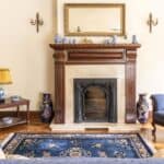 A traditional living room with a fireplace, blue sofas, a blue rug, wooden furniture, vases, and a large mirror above the mantel.