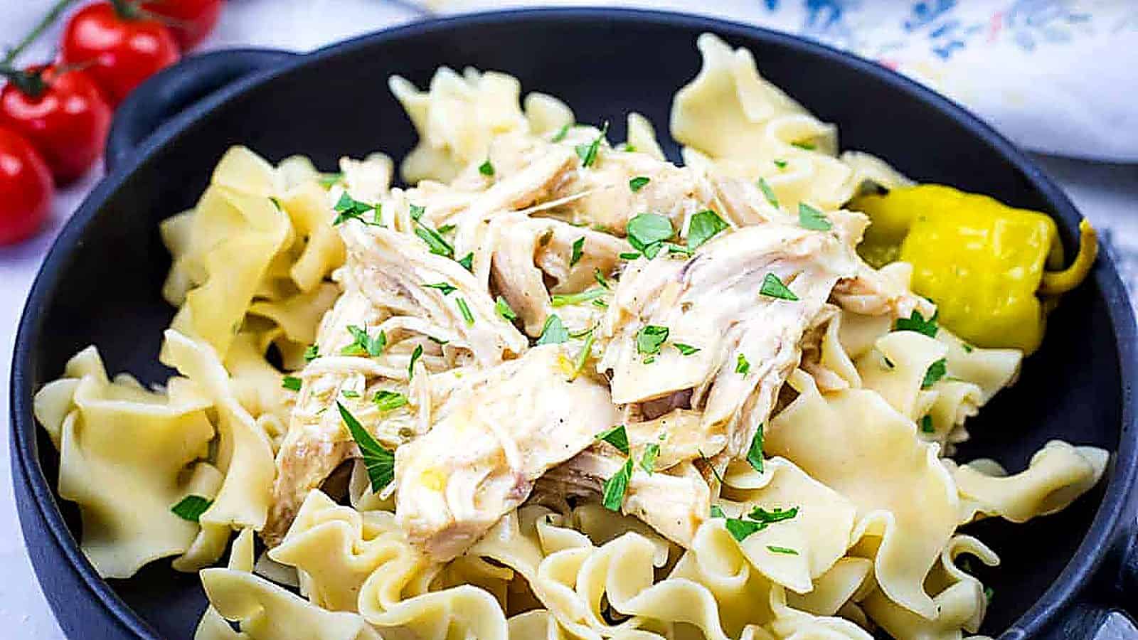 A black bowl containing egg noodles topped with Slow Cooker Mississippi Chicken and garnished with parsley, accompanied by a pickled pepper.