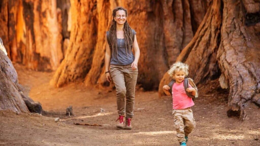 An adult and a child walk and run on a dirt path among large trees in a forest, both smiling and appearing relaxed, celebrating National Park and Recreation Month.