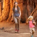 An adult and a child walk and run on a dirt path among large trees in a forest, both smiling and appearing relaxed, celebrating National Park and Recreation Month.