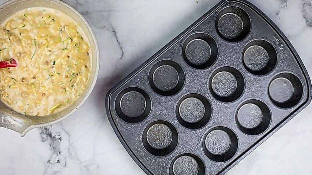A bowl of muffin batter with a red spatula rests beside an empty 12-cup muffin tin on a marble countertop, ready to be filled and baked in your favorite baking pans.