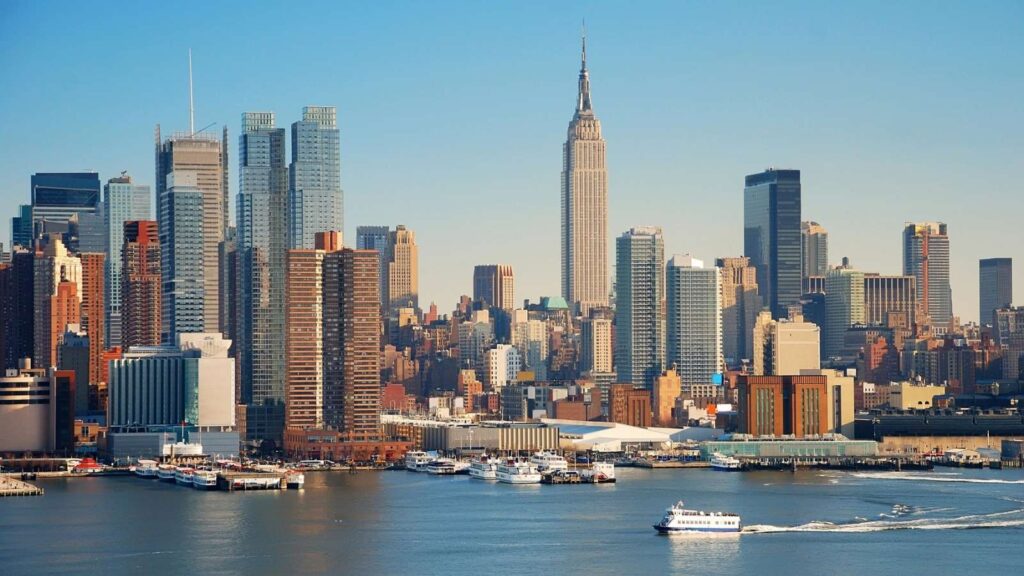 New York City skyline with the Empire State Building, high-rise buildings, and a ferry on the Hudson River under a clear blue sky.