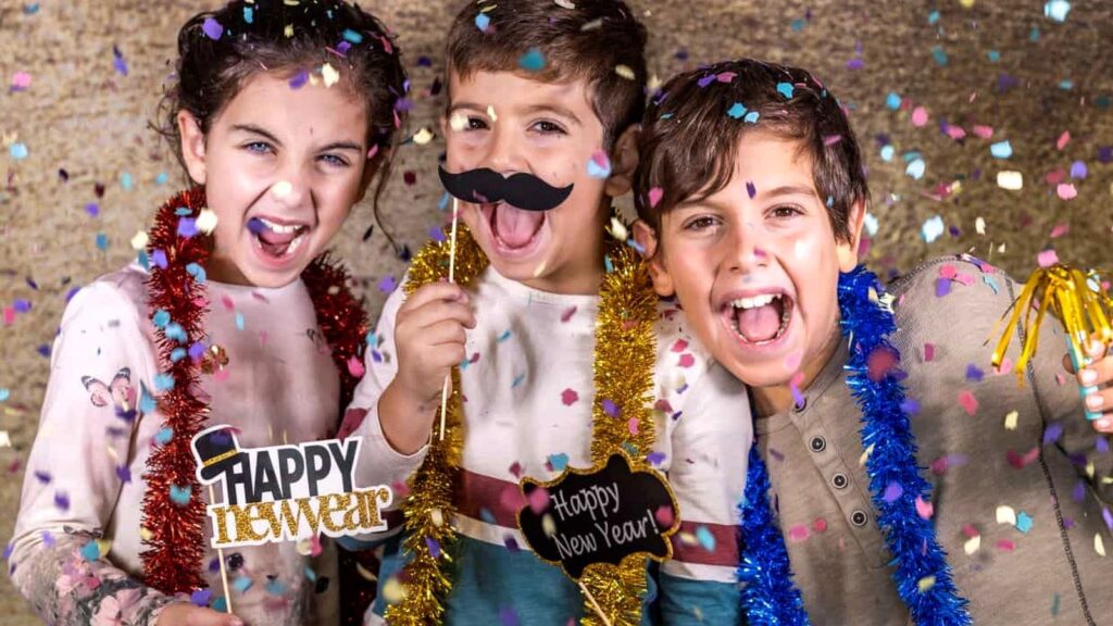 Three children wearing festive tinsel and holding "Happy New Year" props smile and celebrate amid falling confetti.