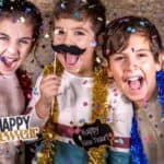 Three children wearing festive tinsel and holding "Happy New Year" props smile and celebrate amid falling confetti.