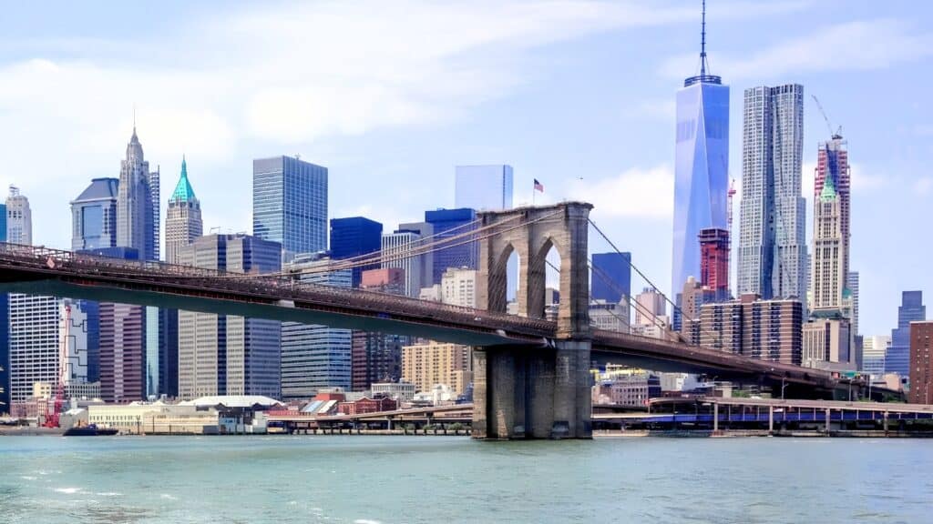 Brooklyn Bridge spans the East River with the Manhattan skyline and One World Trade Center visible in the background on a clear day.