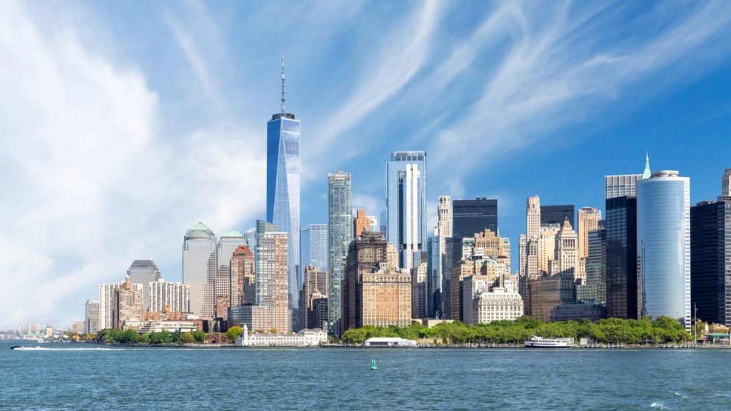 The New York City skyline with tall skyscrapers, including One World Trade Center, viewed from across the water under a partly cloudy sky.