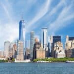 The New York City skyline with tall skyscrapers, including One World Trade Center, viewed from across the water under a partly cloudy sky.